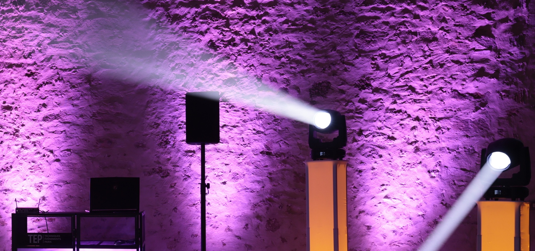 Mise en lumière événementielle professionnelle : deux lyres asservies sur totems rétroéclairés projetant des faisceaux blancs nets contre un mur de pierre ancienne illuminé en violet, avec une enceinte sur pied et un pupitre DJ Techniques Événements Paris (TEP).